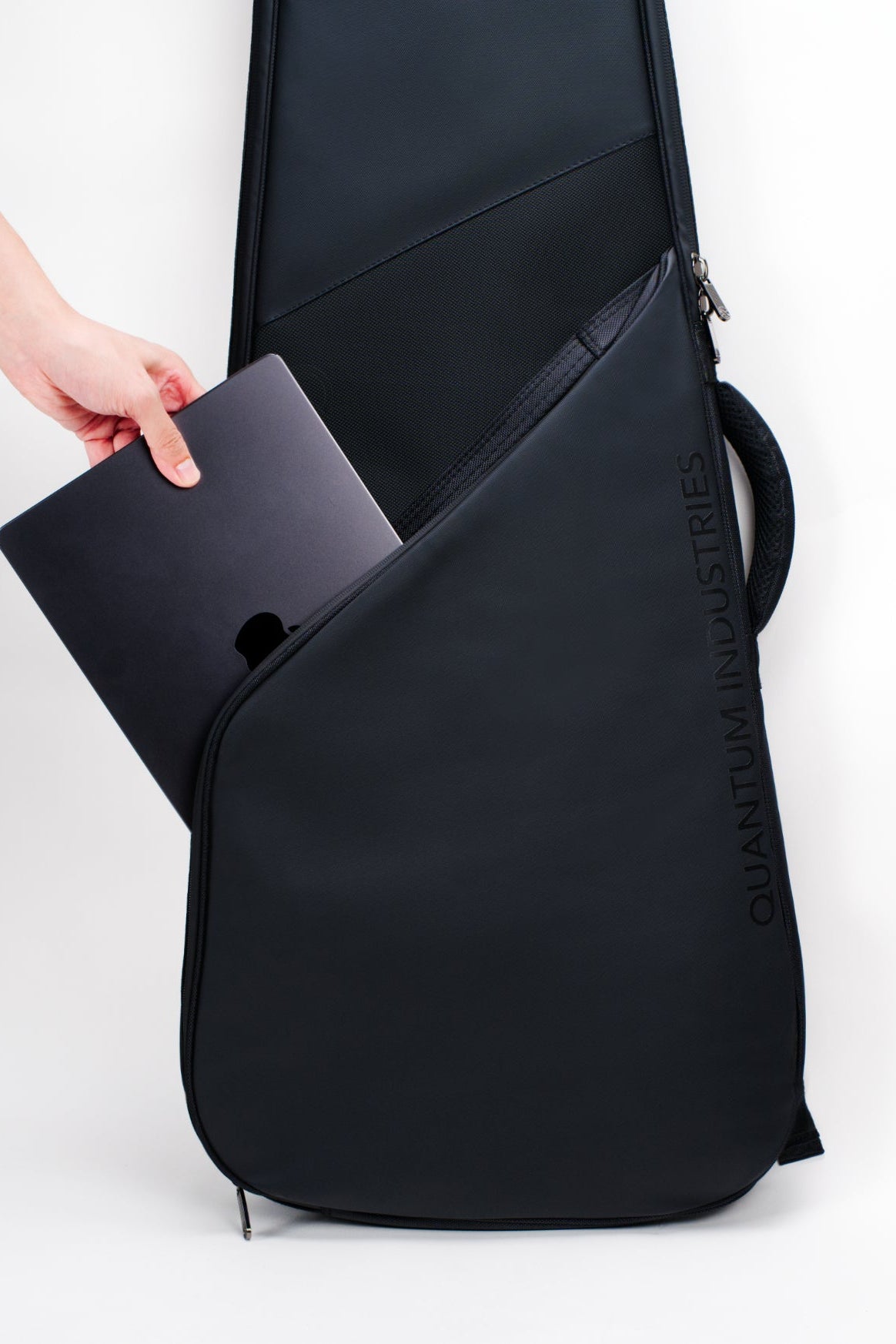 A black guitar gig bag with a hand placing a black laptop into its side laptop compartment, shown against a plain white studio background.