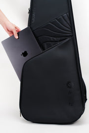 A black laptop backpack shown on a white studio background, with a hand inserting a dark gray laptop into its side pocket. The backpack features a sleek, modern design with a subtle wave pattern on the upper section and a circular logo near the bottom.